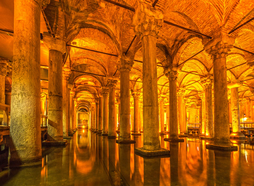 Ancient underground chamber with numerous stone columns and arched ceilings, illuminated by warm golden lights, reflecting on water.